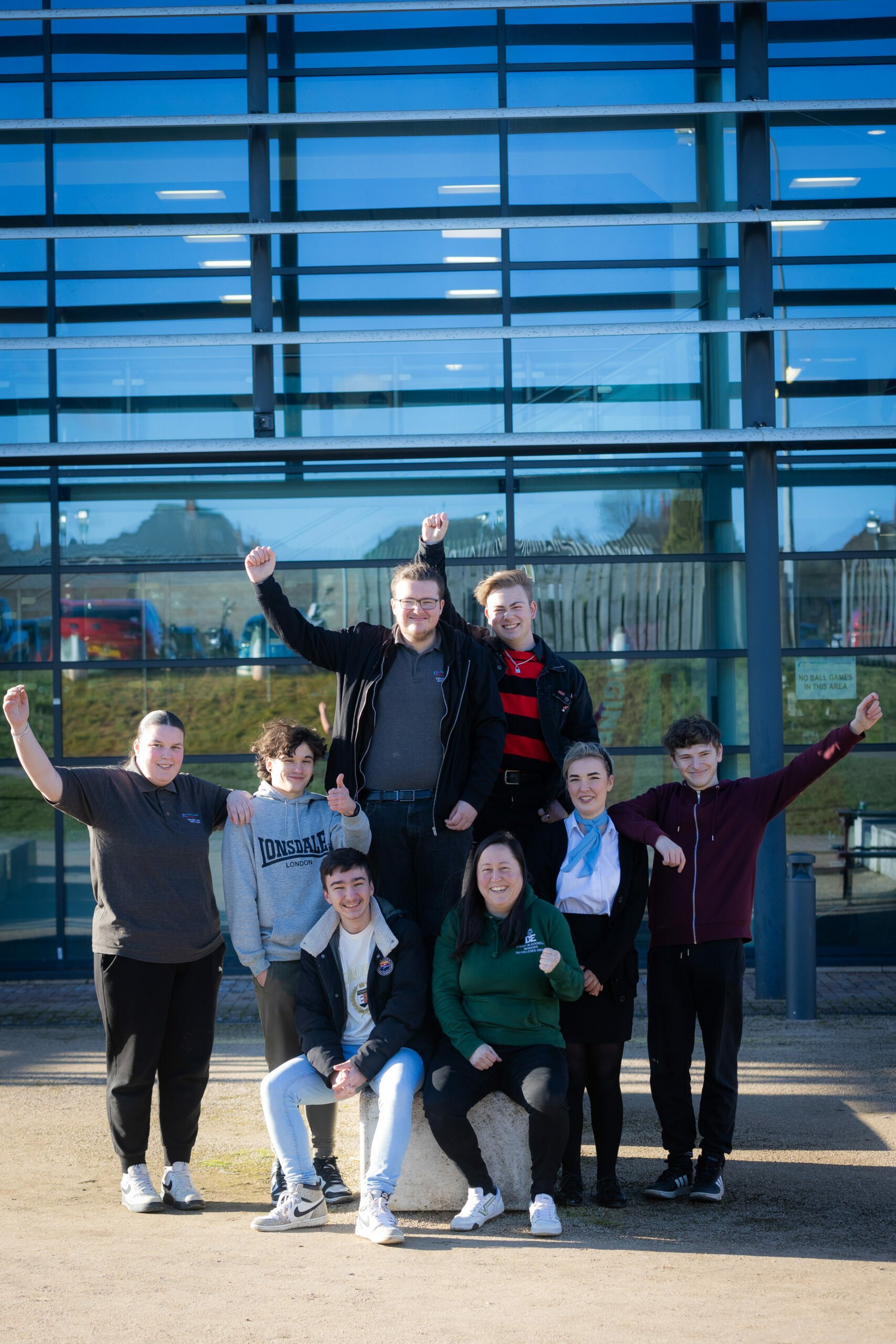 A group of eight young people poses outside a modern building with large glass windows. They are arranged in two rows, with some standing and others sitting on a stone. Everyone is smiling and raising their fists in a celebratory gesture. The background features a grassy area and playground equipment. The scene conveys a sense of joy and camaraderie.