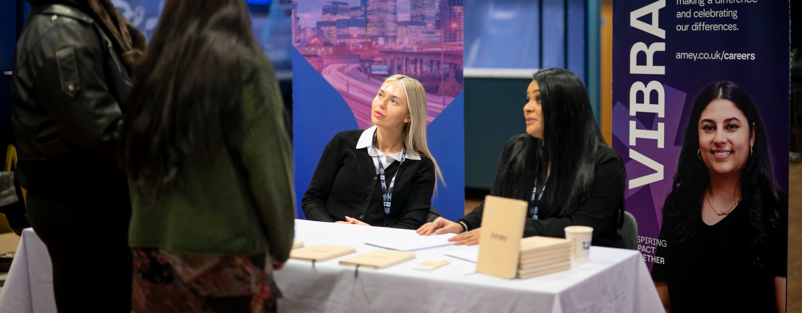 Two women sit at a table with banners for Amey and Vibrant, engaging with two individuals standing in front of them. The setting appears to be a career fair or informational event, with a modern backdrop featuring cityscapes. The women are dressed professionally and are discussing opportunities with the attendees.