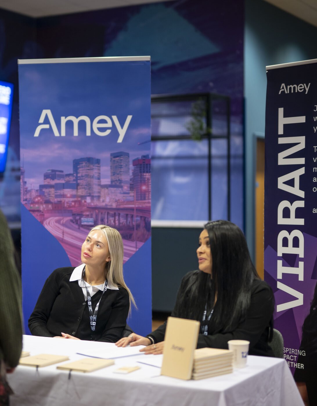 Two women sit at a table with banners for Amey and Vibrant, engaging with two individuals standing in front of them. The setting appears to be a career fair or informational event, with a modern backdrop featuring cityscapes. The women are dressed professionally and are discussing opportunities with the attendees.