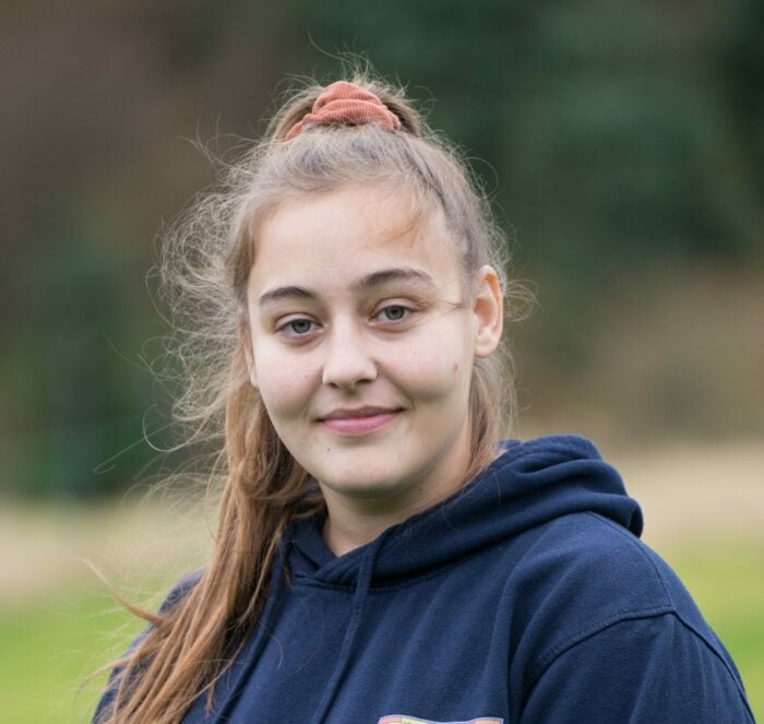 Sophie is standing outdoors, smiling softly at the camera. She is wearing a navy hoodie with a colourful