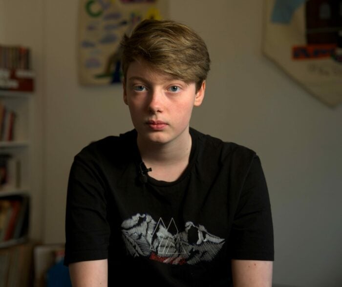 Ellesha has short light-brown hair and a black T-shirt, sits indoors in front of blurred bookshelves, looking calmly at the camera.