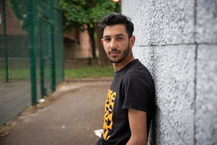 Alyas with short, dark, wavy hair and a trimmed beard stands outdoors, leaning against a textured grey wall. He is wearing a black t-shirt with an orange graphic design and looking directly at the camera with a calm expression. The background includes a pathway, green fencing, grass, and trees.