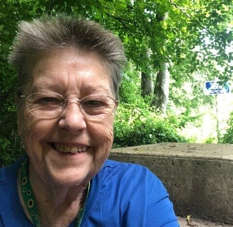 Jean, an older woman with short hair and glasses, smiles warmly in a selfie taken outdoors. She is wearing a blue shirt and a green lanyard, surrounded by lush greenery and trees, with a concrete structure and a partially visible sign in the background