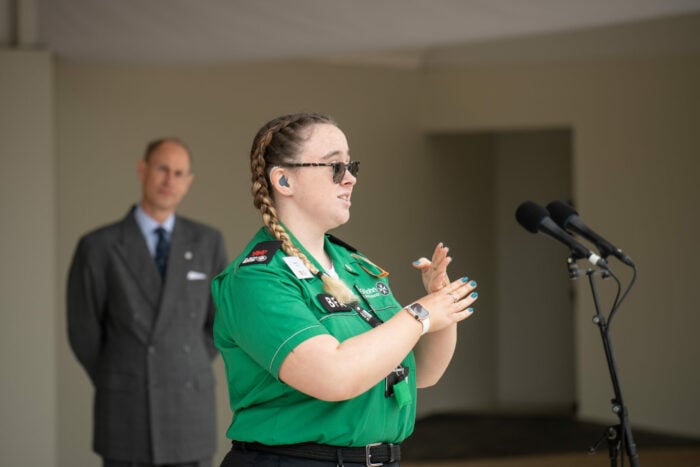 Hafwen, a young woman in a green St John Ambulance uniform, stands at a microphone signing in British Sign Language. She wears glasses, has braided hair, and an Apple Watch on her wrist. A man in a suit stands in the background watching attentively.