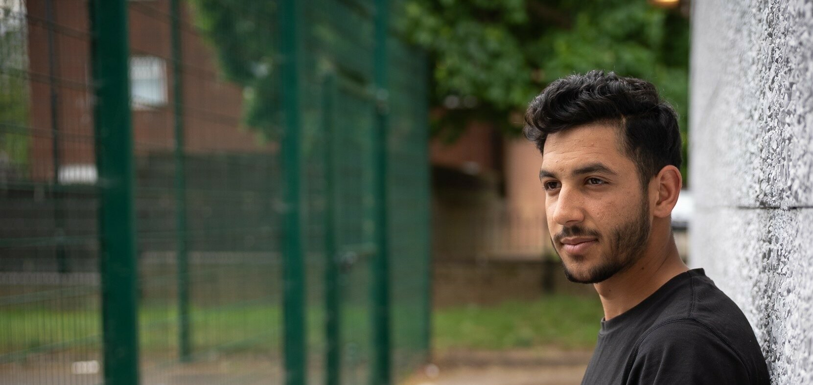 Alyas with short, dark, wavy hair and a trimmed beard stands outdoors, leaning against a textured grey wall. He is wearing a black t-shirt with an orange graphic design and looking directly at the camera with a calm expression. The background includes a pathway, green fencing, grass, and trees.