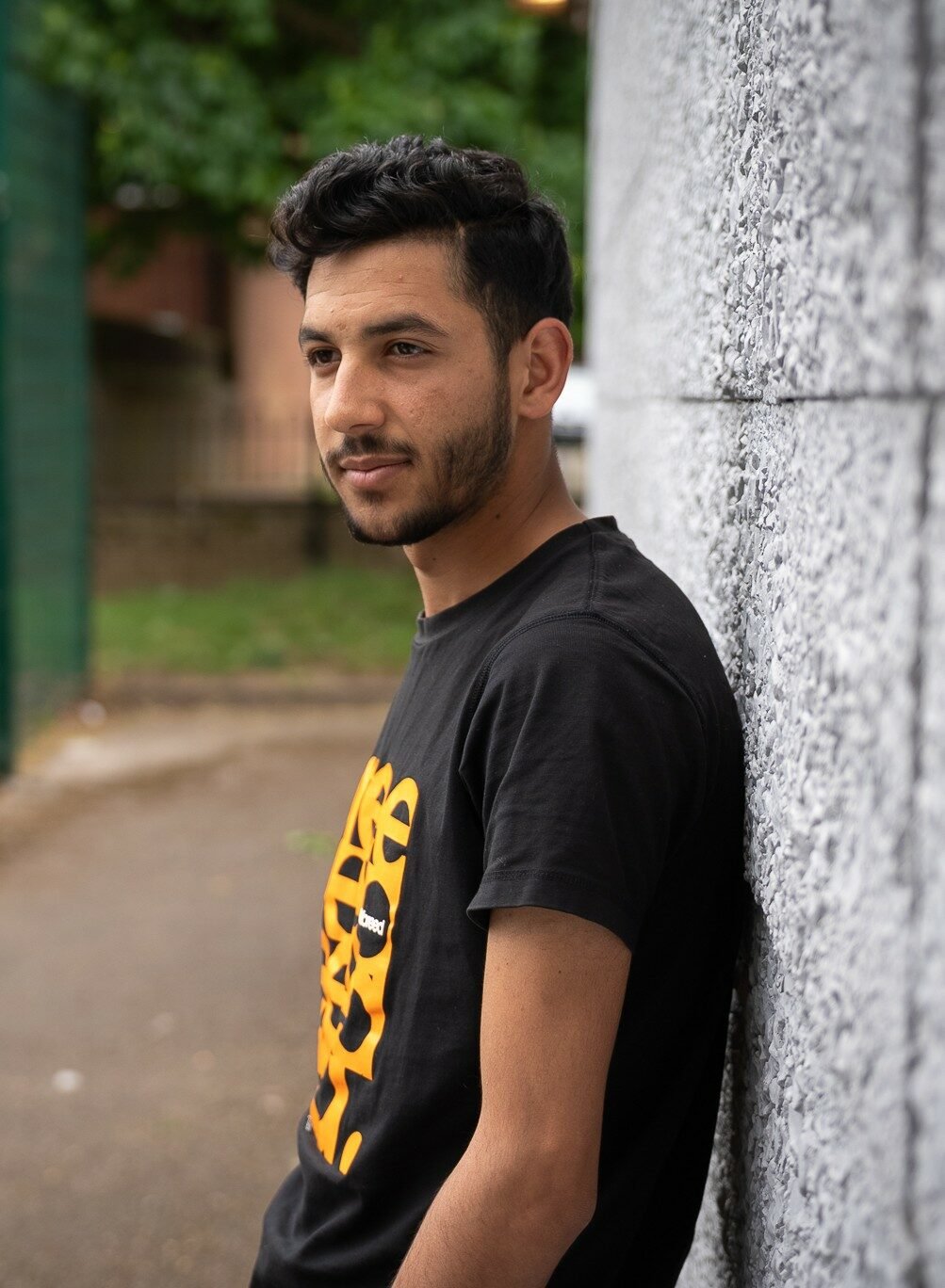 Alyas with short, dark, wavy hair and a trimmed beard stands outdoors, leaning against a textured grey wall. He is wearing a black t-shirt with an orange graphic design and looking directly at the camera with a calm expression. The background includes a pathway, green fencing, grass, and trees.