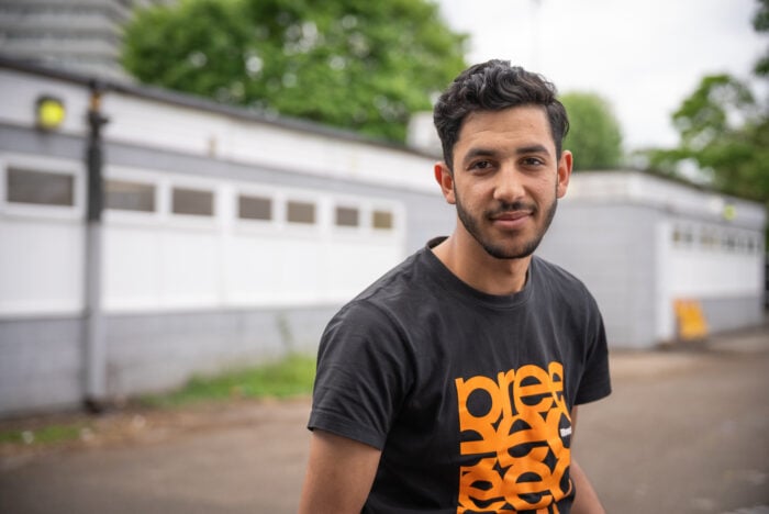 Alyas, a young man with short dark hair and a trimmed beard, wearing a black t-shirt with bold orange text, standing outdoors in front of a light gray building with white panels and windows. Trees and a paved area are visible in the background.