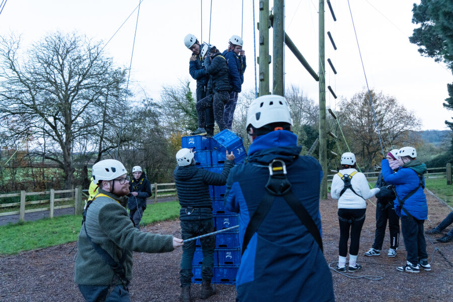 Three people are making a crate tower together, supported by people belaying and passing on crates on the ground.
