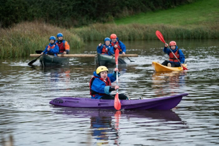 A group of six people kayaking on a calm body of water surrounded by green grass and reeds along the water's edge.