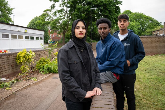 Three young individuals stand together outdoors. The person in the foreground, a woman wearing a black hijab and a dark jacket, looks confidently at the camera. Behind her, two young men are positioned slightly to the left and right. One man is wearing a blue jacket and has a serious expression, while the other is dressed in a dark hoodie and appears contemplative. The background features greenery and a low brick wall, suggesting a community setting.