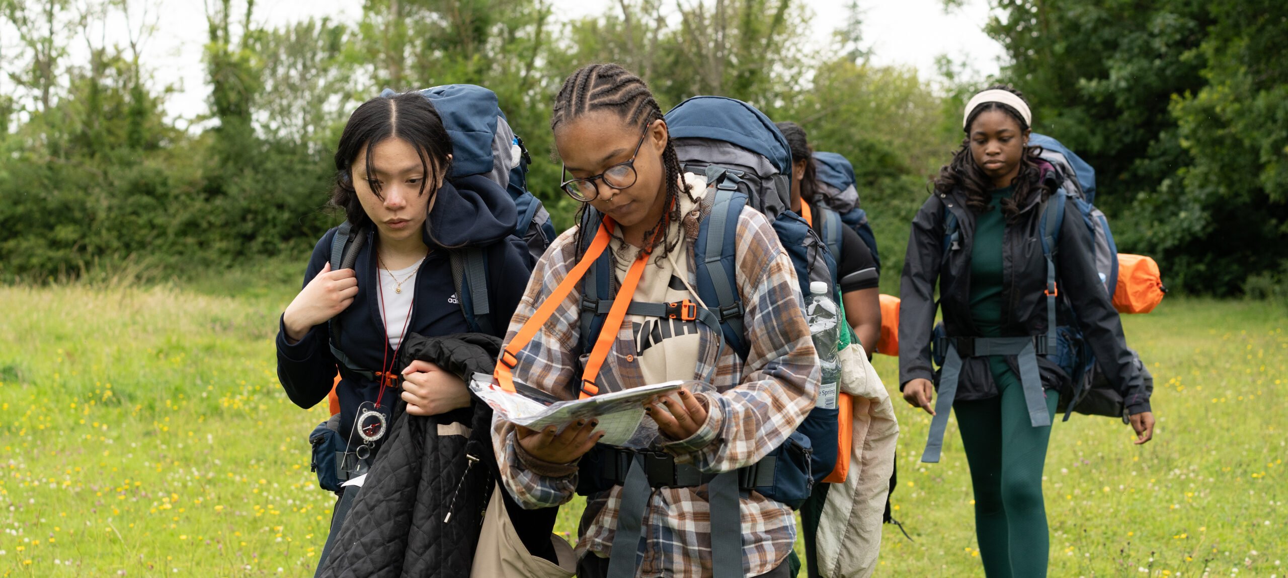 A group of four young hikers is walking through a grassy field. One person, wearing glasses and a plaid shirt, is reading a map, while another person, dressed in a black jacket, looks concerned. The other two hikers are following behind, carrying large backpacks. The background features trees and a cloudy sky, suggesting an overcast day.