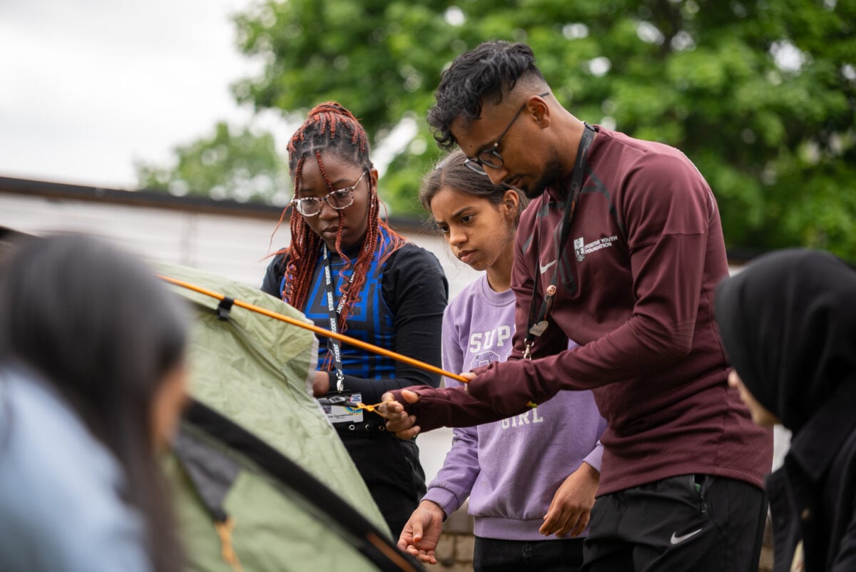 Three individuals are gathered around a tent, with one person adjusting a strap while another observes closely. The group appears engaged in a camping activity, surrounded by greenery in the background.