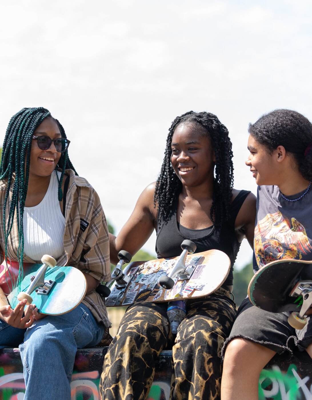 Amaris, Josie and Tilly sitting outdoors in a park-like setting, each holding a skateboard. The background features trees and greenery.