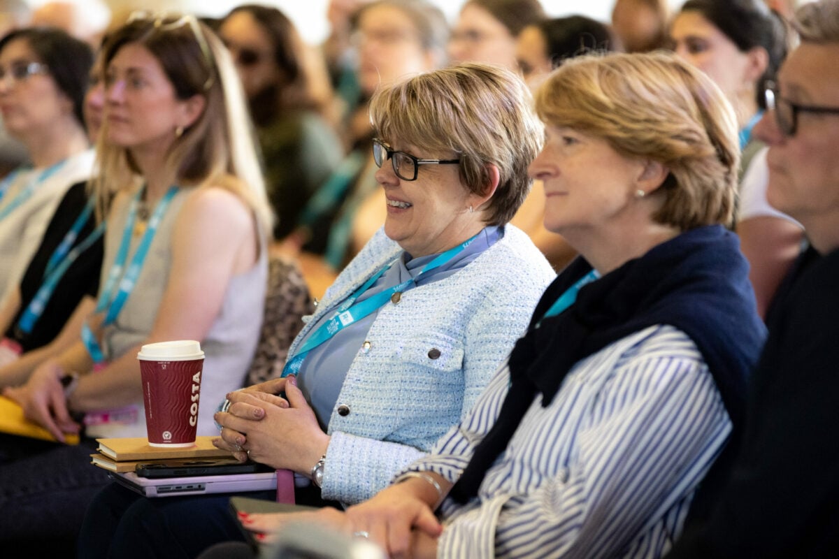 A group of people sitting in an audience, attentively listening to a presentation. In the foreground, two women are smiling; one has short, light brown hair and glasses, wearing a light blue jacket, while the other has medium-length blonde hair and is dressed in a navy sweater. Several attendees in the background are also visible, with a mix of expressions. A coffee cup is placed on the table in front of the woman in the blue jacket.