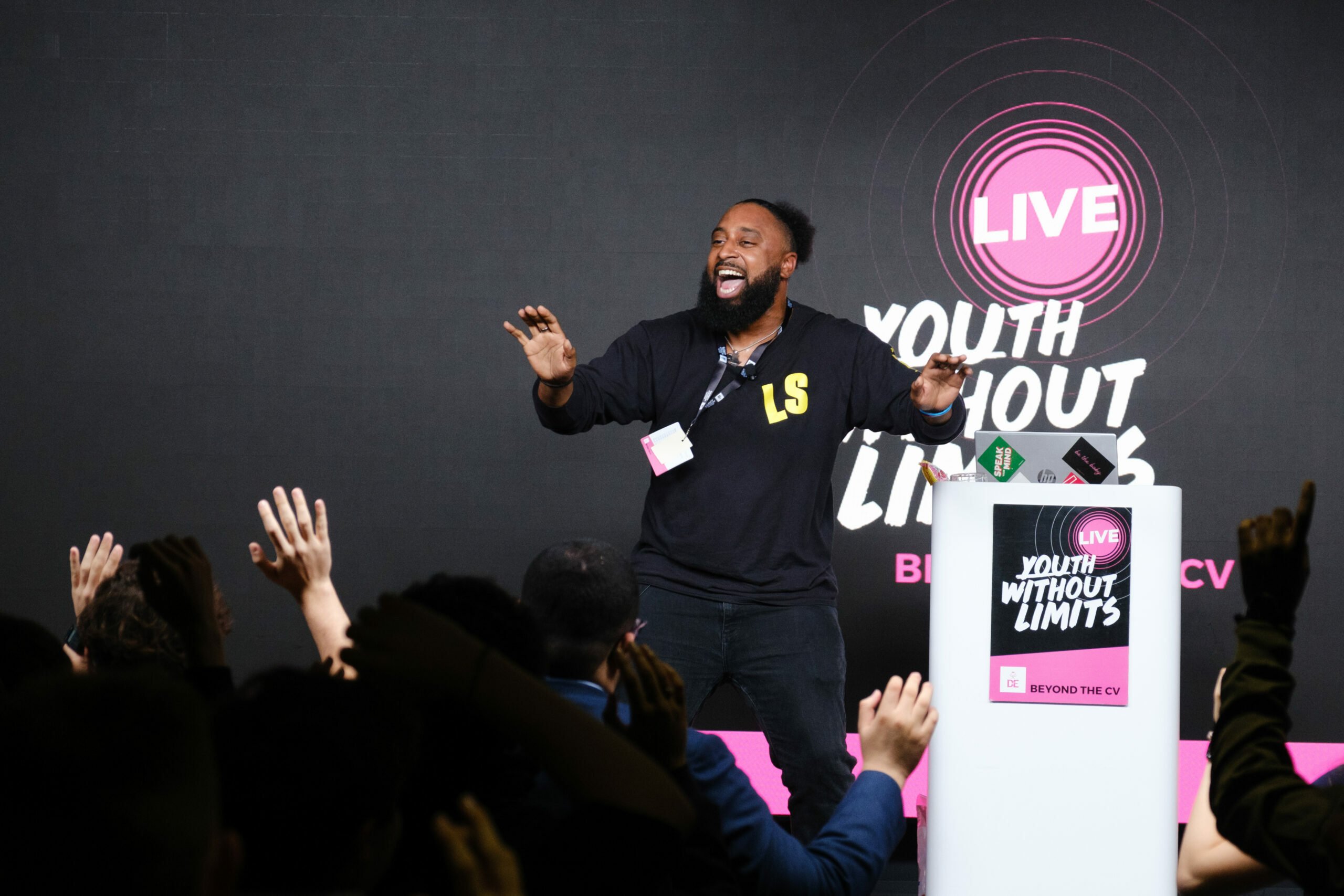A speaker with a beard and wearing a black sweatshirt is energetically engaging with an audience at a live event titled 