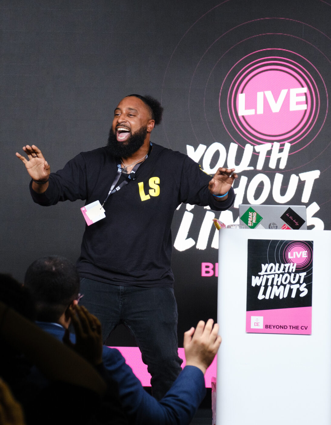 A speaker with a beard and wearing a black sweatshirt is energetically engaging with an audience at a live event titled 