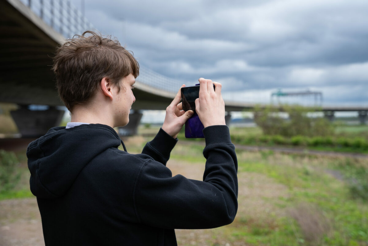 Taylor taking a photo with a smartphone outdoors, with an overpass bridge, greenery, and a cloudy sky in the background.