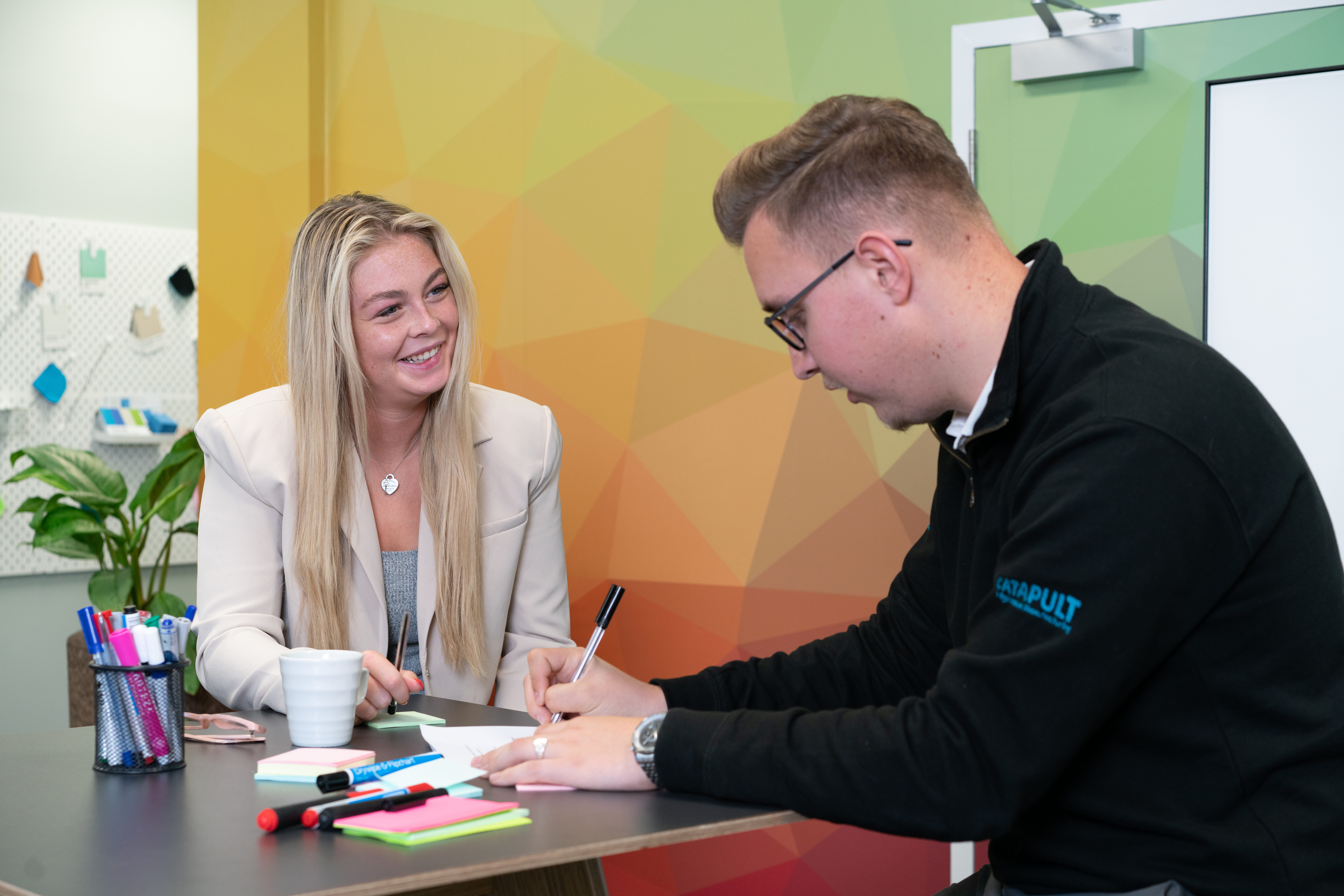 Two people sitting at a table engaged in discussion, with one writing on paper and the other observing. The table is scattered with stationery items including pens, markers, sticky notes, and a white coffee cup. A colorful geometric background and a plant are visible.