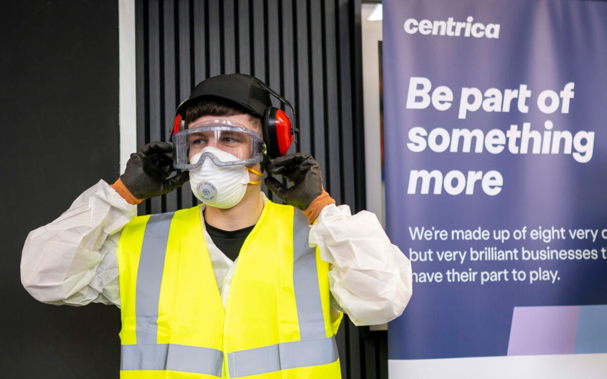 A person wearing a yellow safety vest, protective gloves, a face mask, and safety goggles is adjusting their ear protection. They are standing in front of a banner that reads 