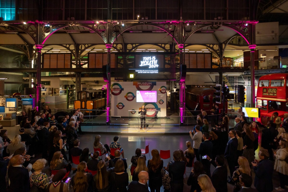 Alt text: A large crowd gathers in a museum space with industrial architecture, featuring colorful lighting. A stage is set up in the center with a screen displaying 