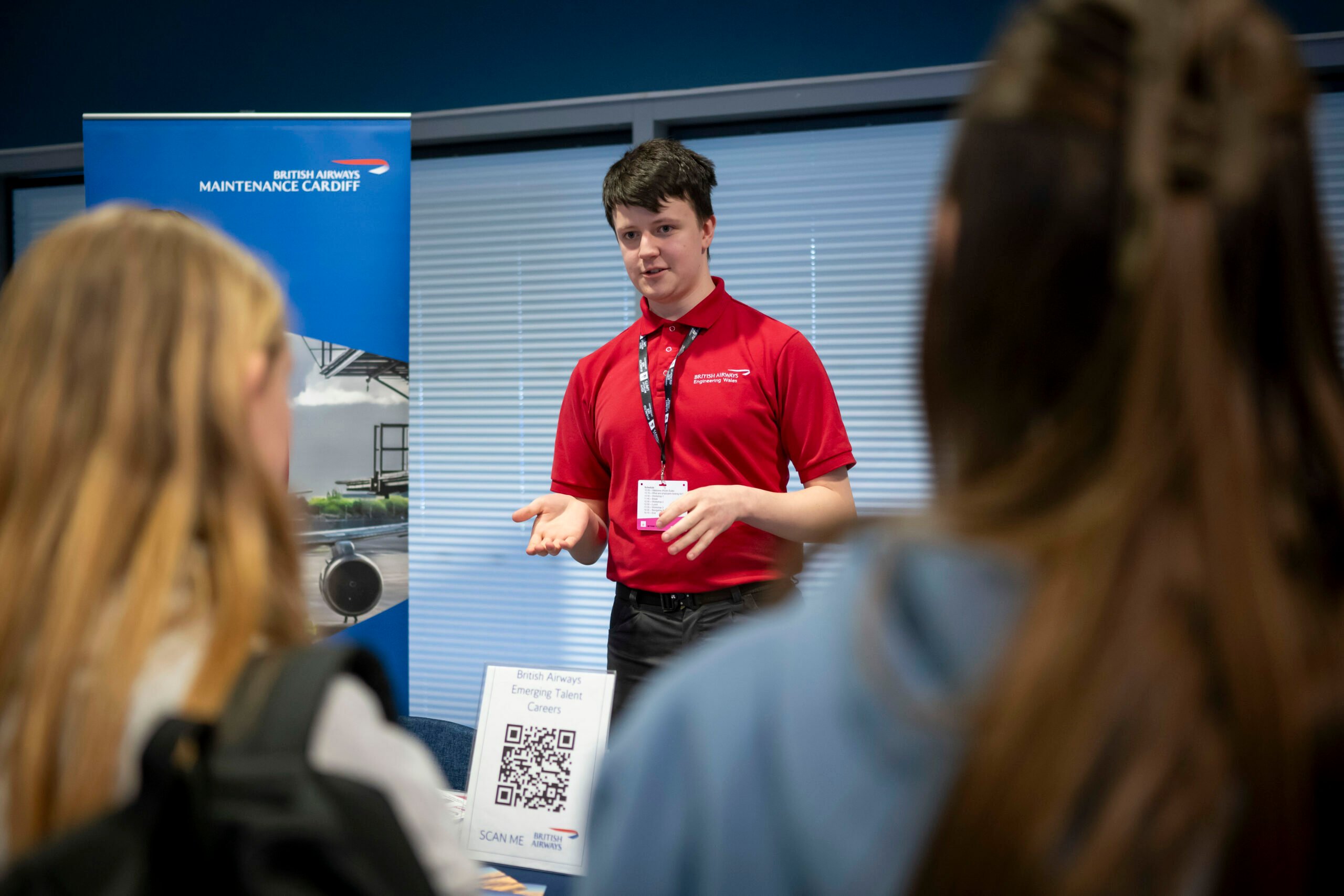 A young man in a red polo shirt stands in front of a display, speaking to two people whose backs are facing the camera. The background features a blue wall and a banner with the text 