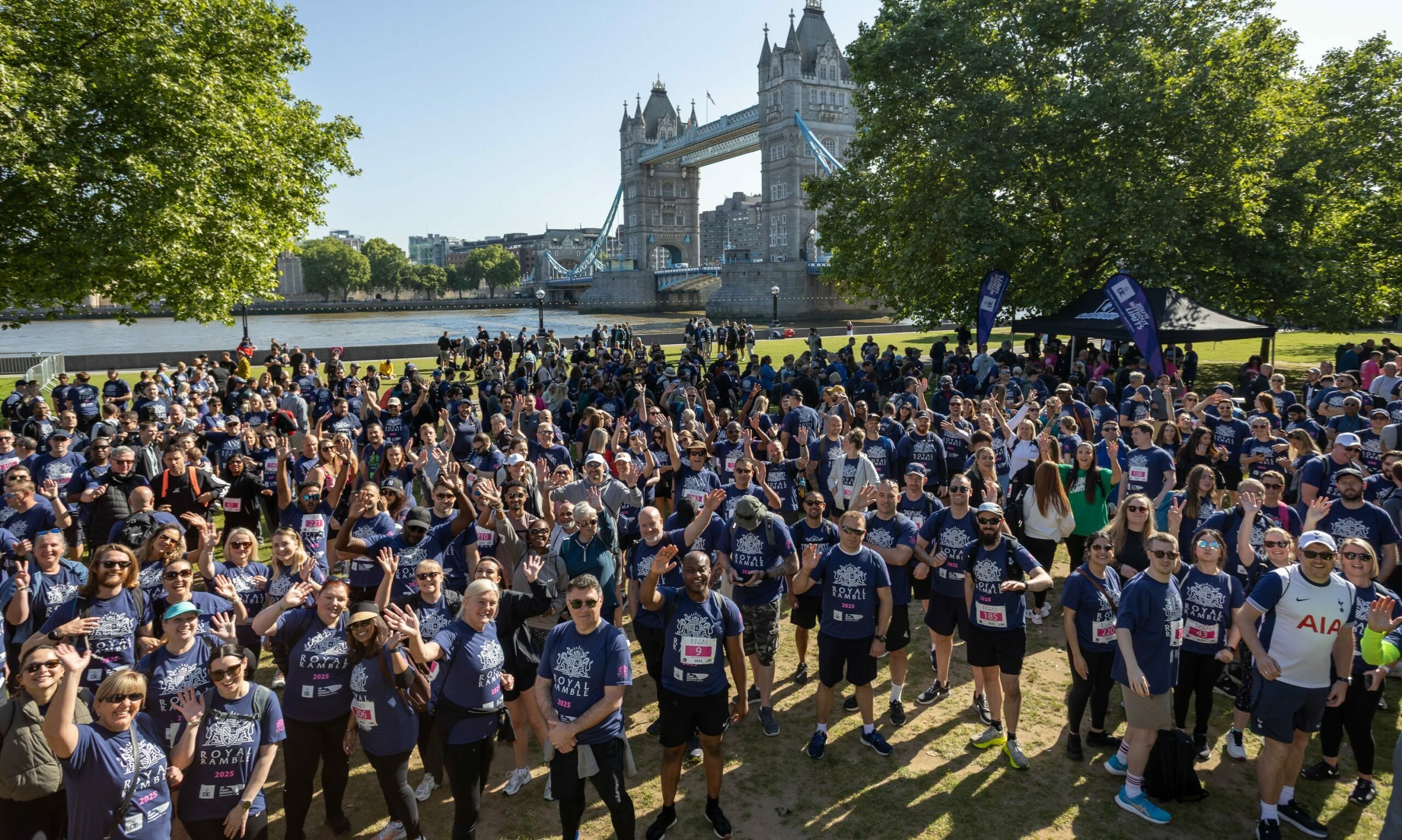 A large crowd of participants wearing matching blue shirts gathers in a park near Tower Bridge in London. The scene is lively, with many people smiling and waving at the camera. The iconic bridge is visible in the background, along with trees and a clear blue sky.