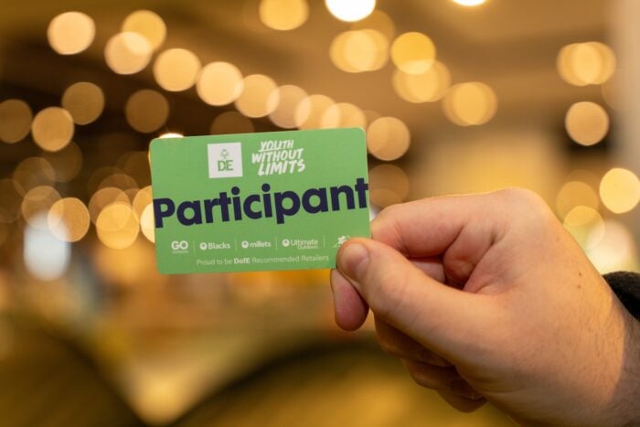 A hand holding a green card labeled 'Participant'. The card features logos and text including 'DE', 'YOUTH WITHOUT LIMITS', 'GO Blacks millets Ultimate Outdoors', and 'Proud to be DofE Recommended Retailers'.