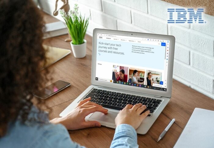 Person working on a laptop displaying the IBM SkillsBuild website at a wooden desk with a notebook, pen, and potted plant; IBM logo visible on a white brick wall in the background.