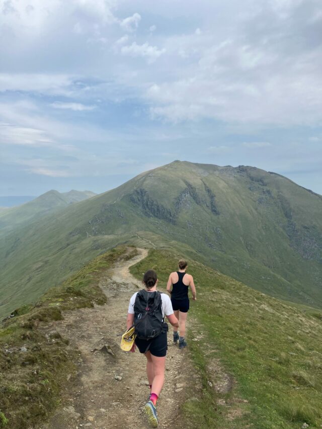 Two people walking on a path on the ridge of a mountain with a view of another mountain ahead.