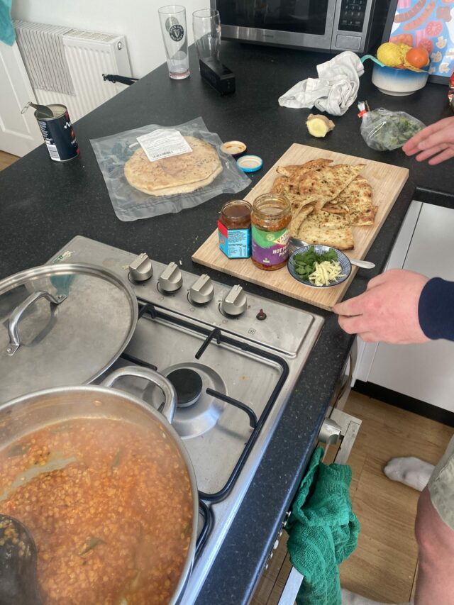 A kitchen counter and hob with a lentil curry cooking and a chopping board with naan and chutneys.
