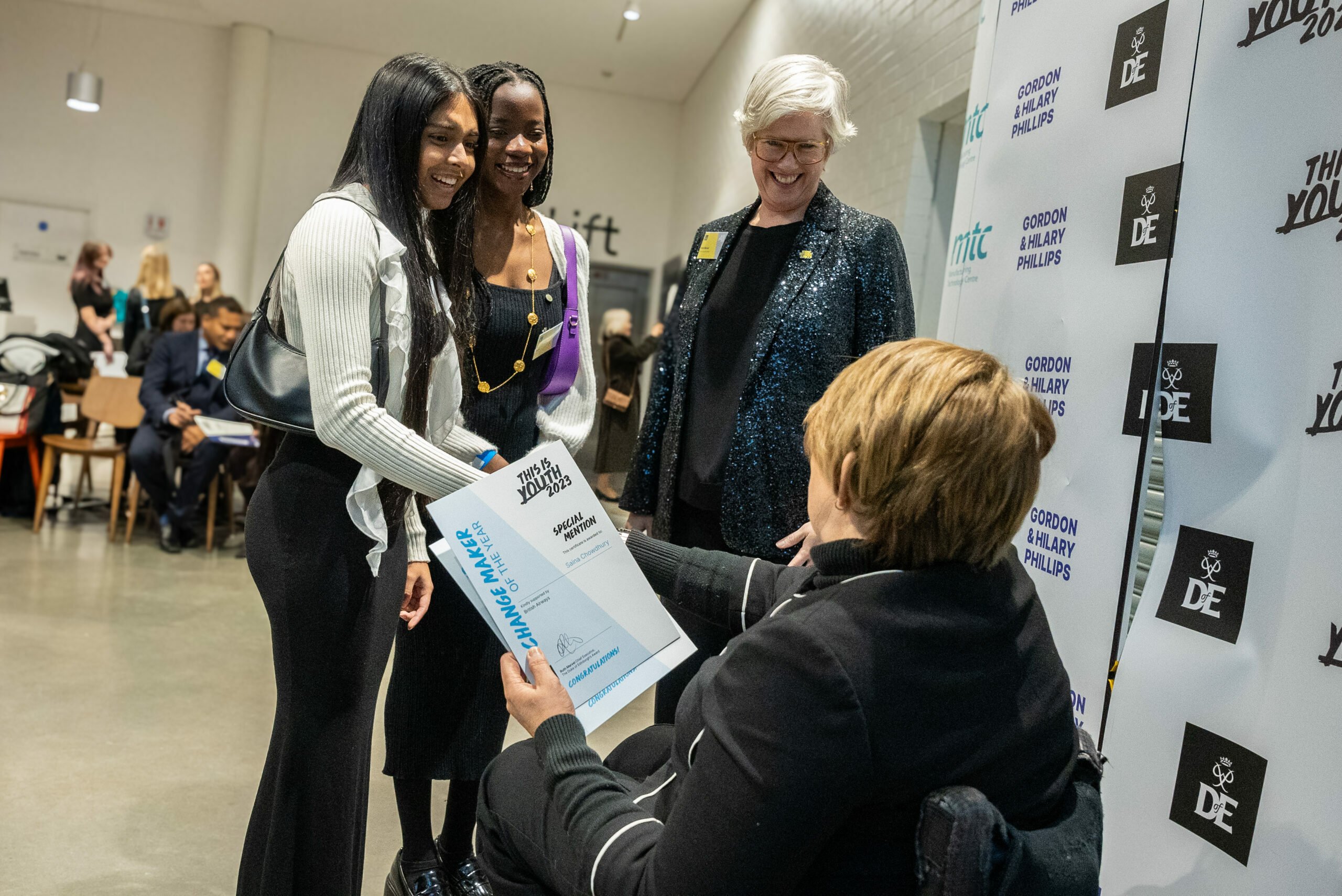 Four individuals at an indoor event, with one seated person holding a certificate that reads 'Gordon & Hillary Phillips' and 'The Young DE 2023'. A banner with logos is visible in the background, indicating an award presentation or recognition ceremony.