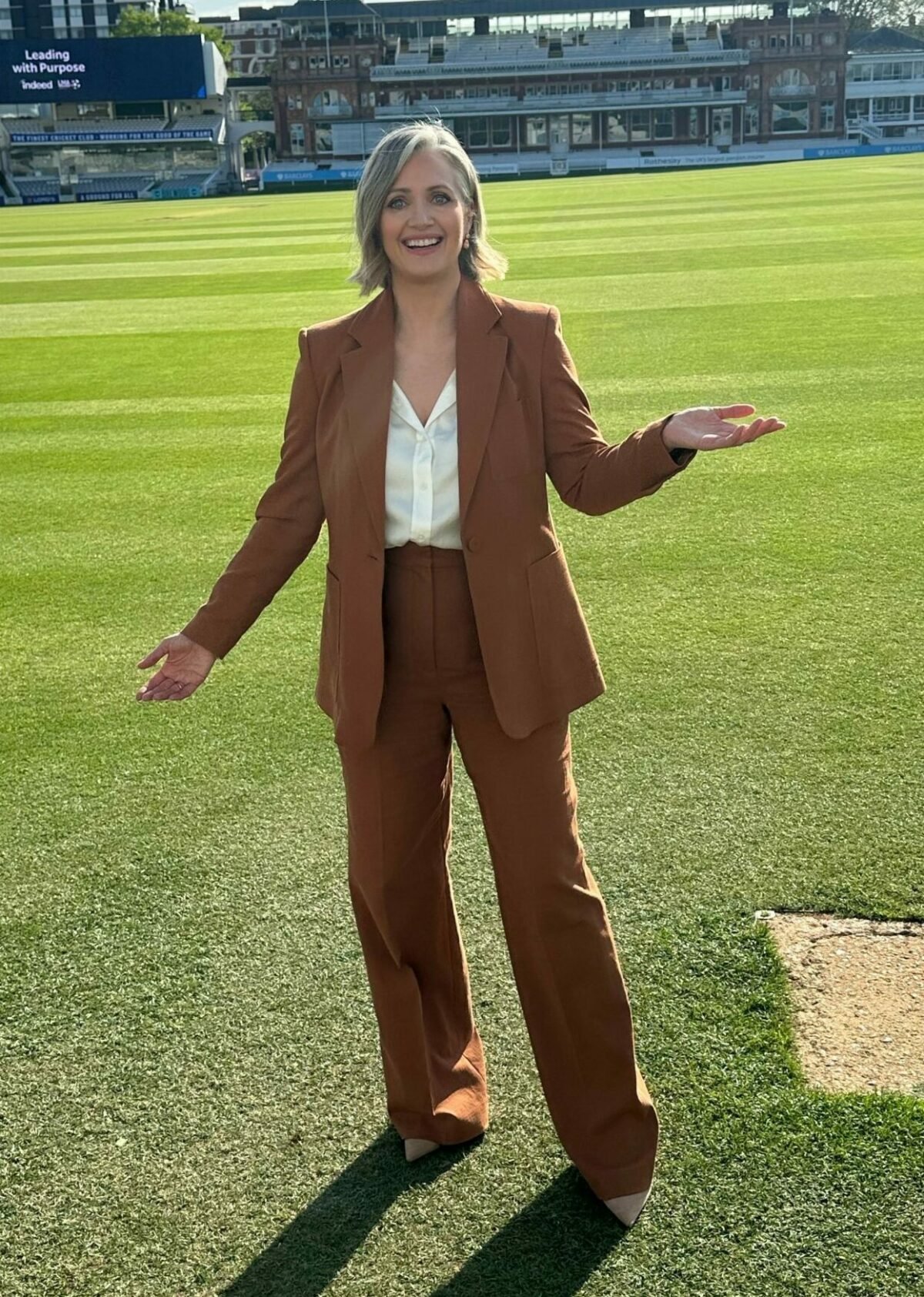 Hayley wearing a brown suit standing on a football pitch holding her arms out to the side.