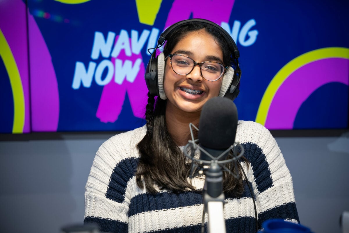 Abiya wearing glasses and headphones speaking into a microphone in a recording studio, with a colorful 'NAVIGATING NOW' sign in the background.