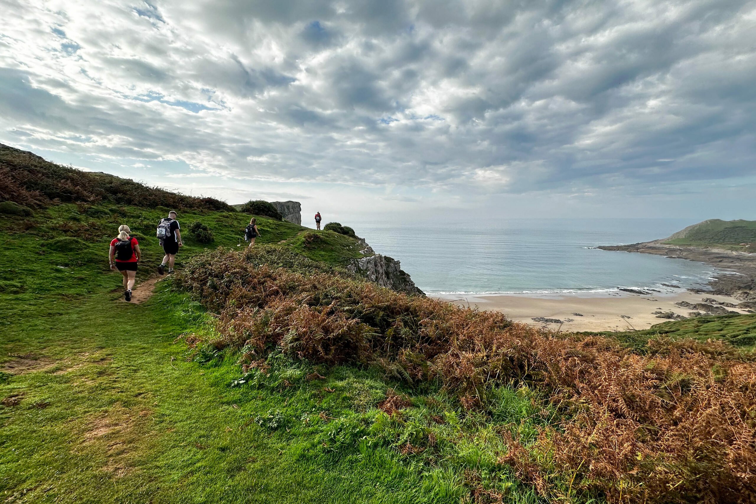 Group of hikers walking along a grassy coastal path near a cliff edge, overlooking a calm ocean under a partly cloudy sky, with lush greenery and sandy beach below.