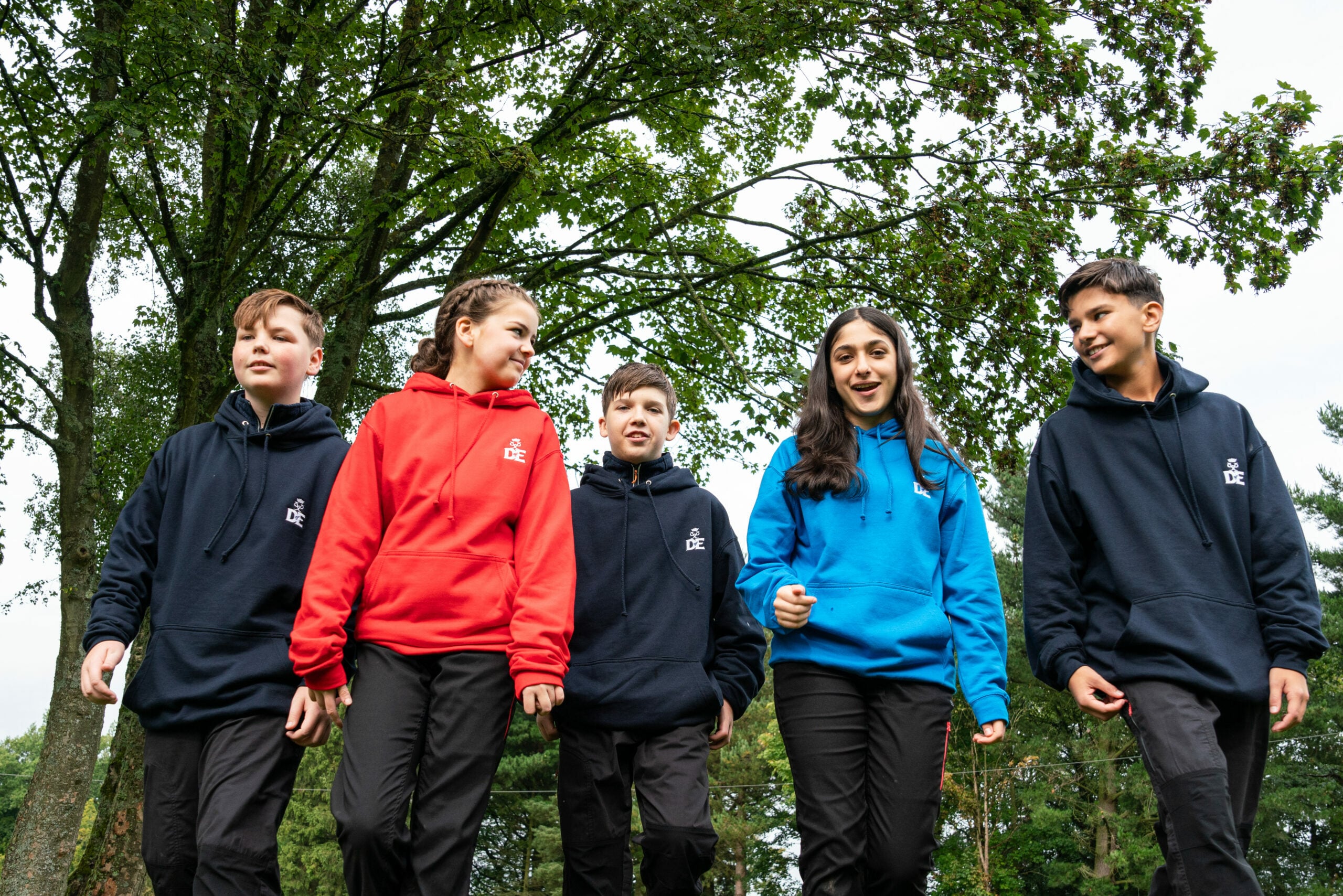 Five individuals walking outdoors on a grassy field with trees in the background. They are wearing black trousers and hoodies in different colours—two in navy blue, one in red, and one in bright blue—with a small DofE logo on the chest. The group appears to be engaged in an outdoor activity or expedition.