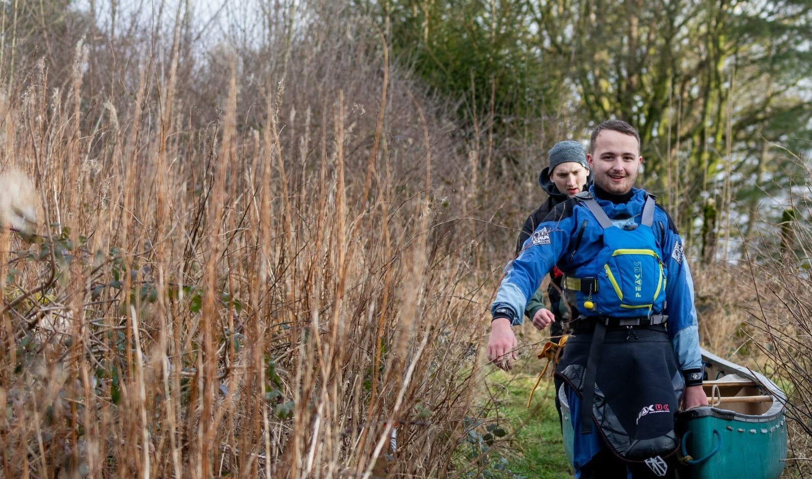 Two individuals wearing blue waterproof gear and life jackets are walking along a narrow grassy path surrounded by tall dry vegetation. They are carrying a green canoe together, with one person holding the front end and the other at the back. The background shows leafless bushes and trees, suggesting a cold or early spring season.