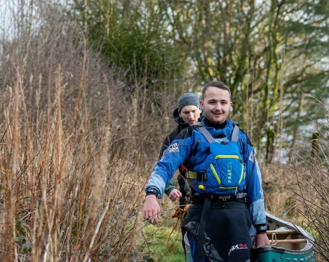 Two individuals wearing blue waterproof gear and life jackets are walking along a narrow grassy path surrounded by tall dry vegetation. They are carrying a green canoe together, with one person holding the front end and the other at the back. The background shows leafless bushes and trees, suggesting a cold or early spring season.