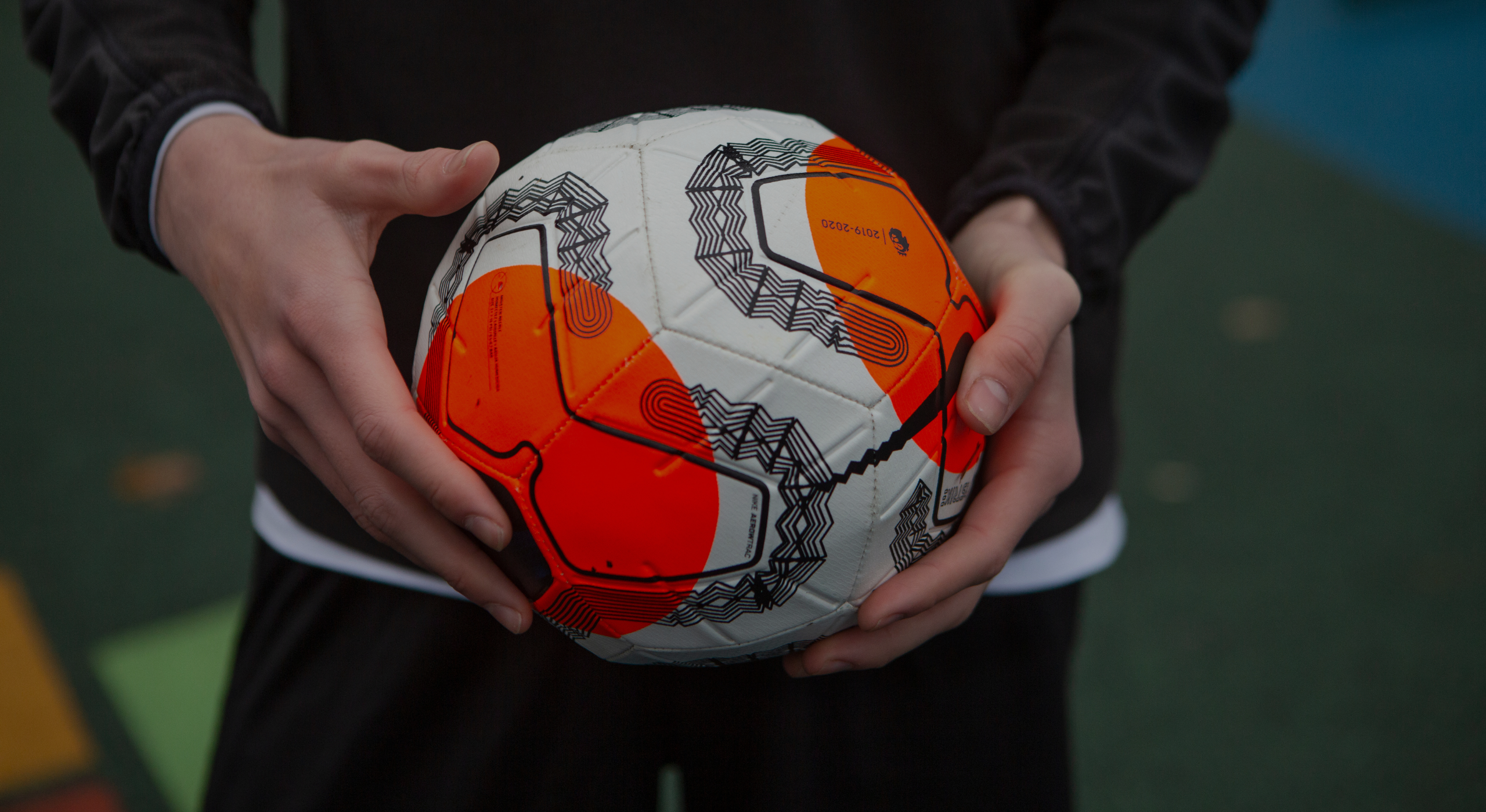Person holding a white, orange, and black geometric-patterned soccer ball on an outdoor sports court with colorful ground markings.