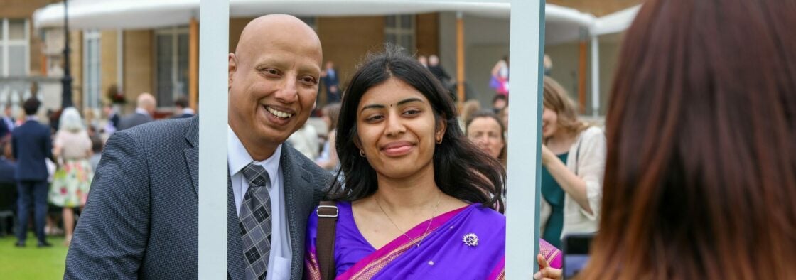 A father in a suit and daughter in a sari standing in a D of E frame in front of Buckingham Palace.
