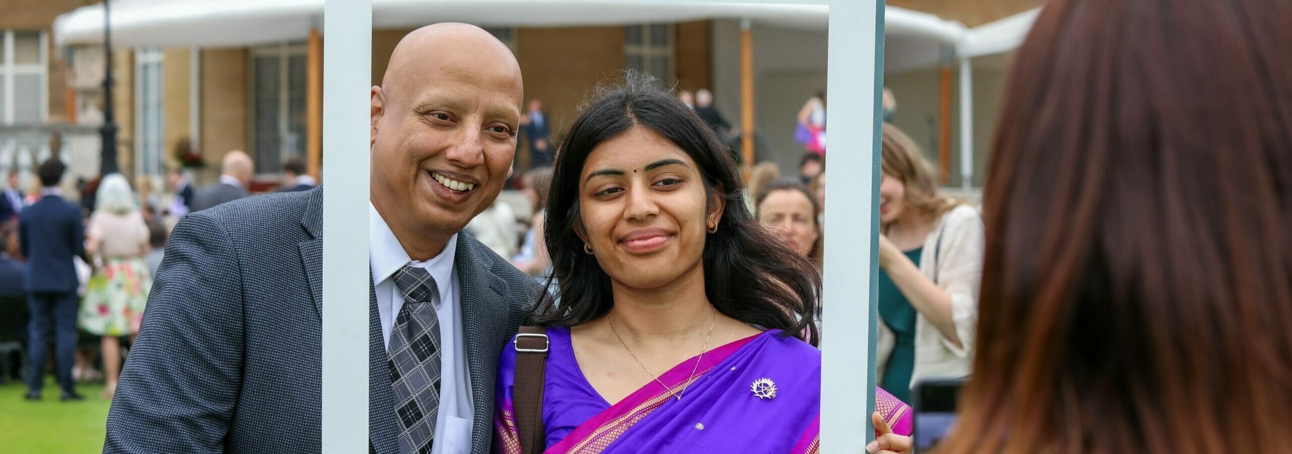 A father in a suit and daughter in a sari standing in a D of E frame in front of Buckingham Palace.