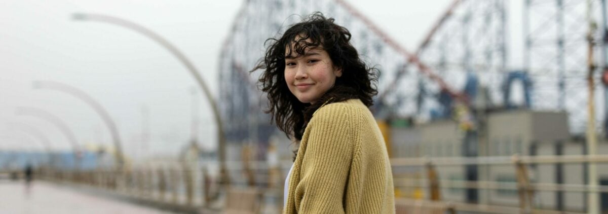 A young person with curly black hair and a yellow cardigan smiling looking over their shoulder.