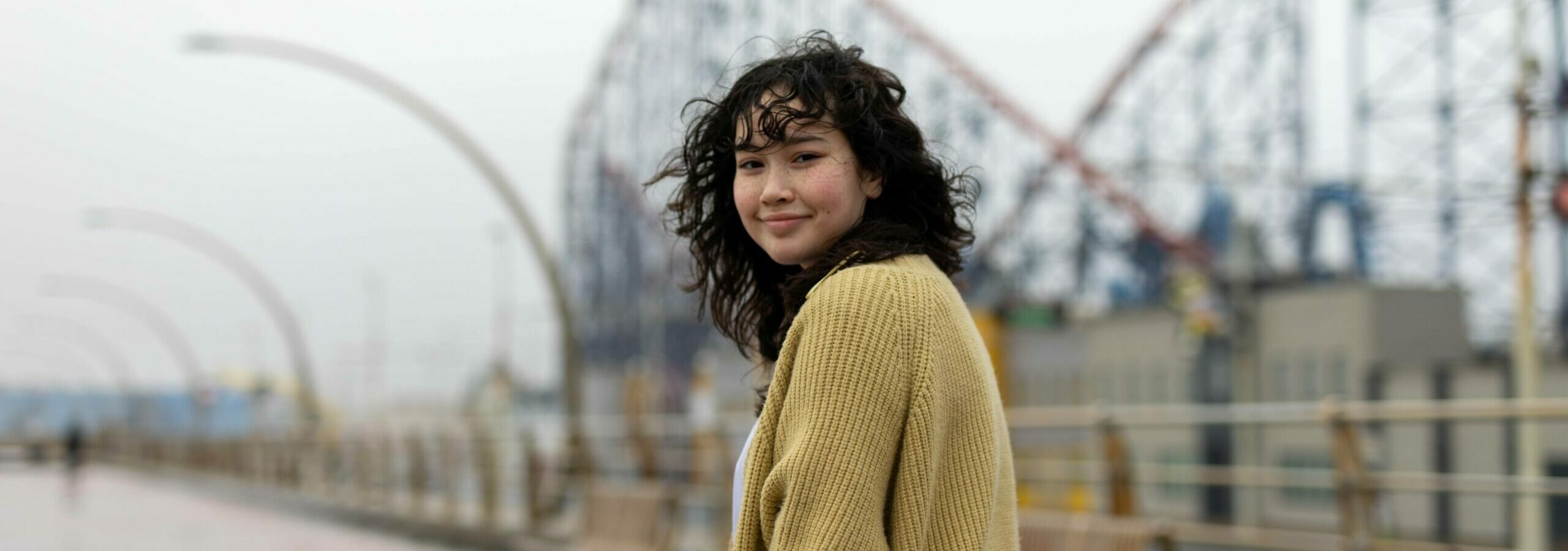 A young person with curly black hair and a yellow cardigan smiling looking over their shoulder.