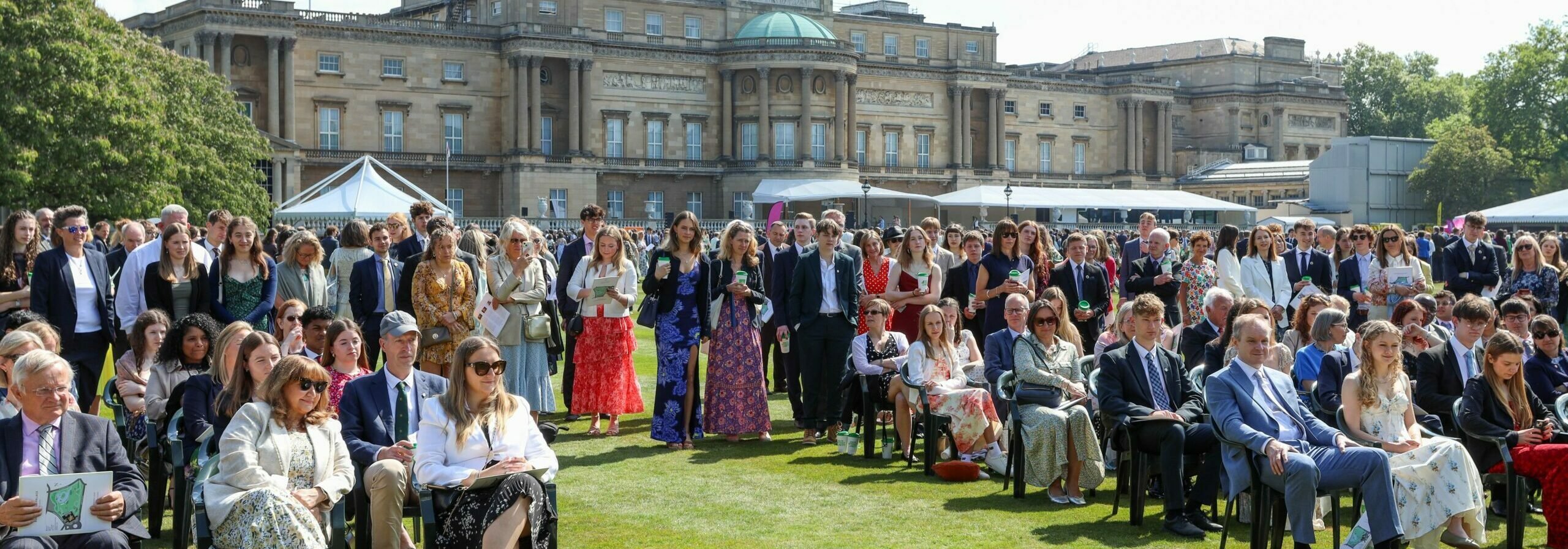 A crowd of people sitting and standing in Buckingham Palace gardens in the sun.