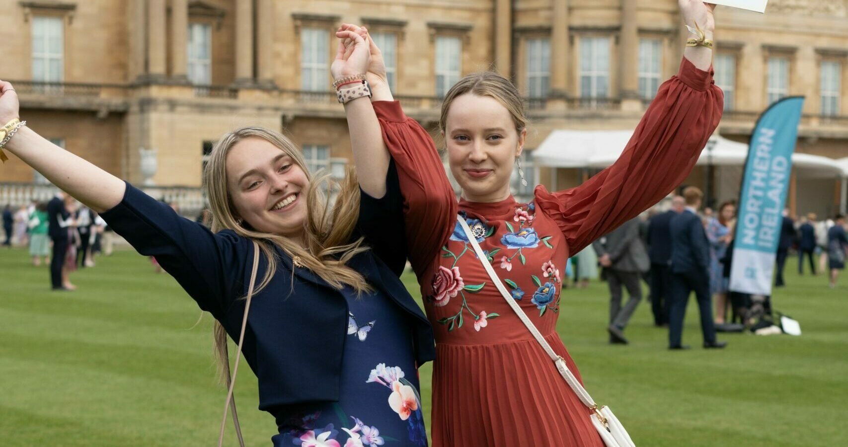 Two young people wearing dresses holding their hands in the air to celebrate in front of Buckingham Palace.
