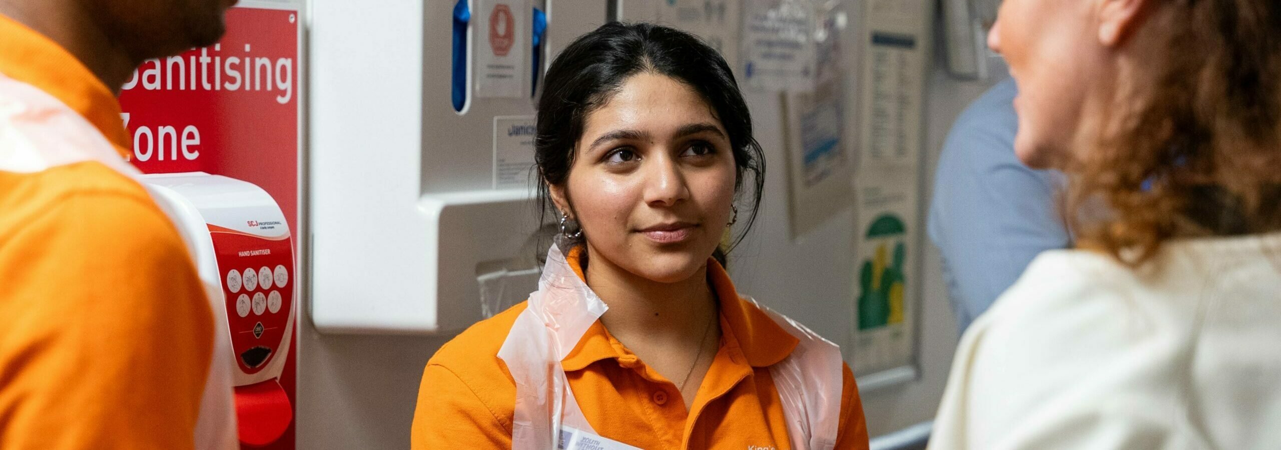A D of E volunteer in a orange tshirt and plastic apron in a hospital.