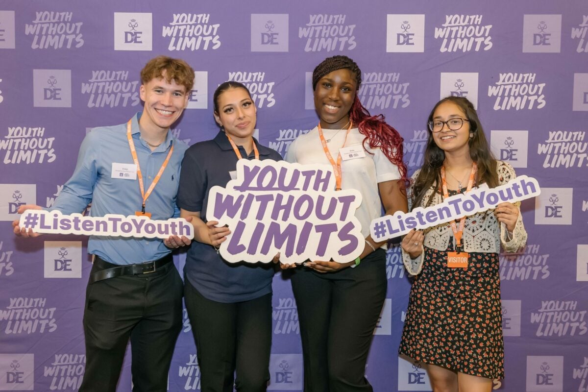 Four D of E Youth Ambassadors standing in front of a purple wall holding signs that say listen to youth and Youth Without Limits.