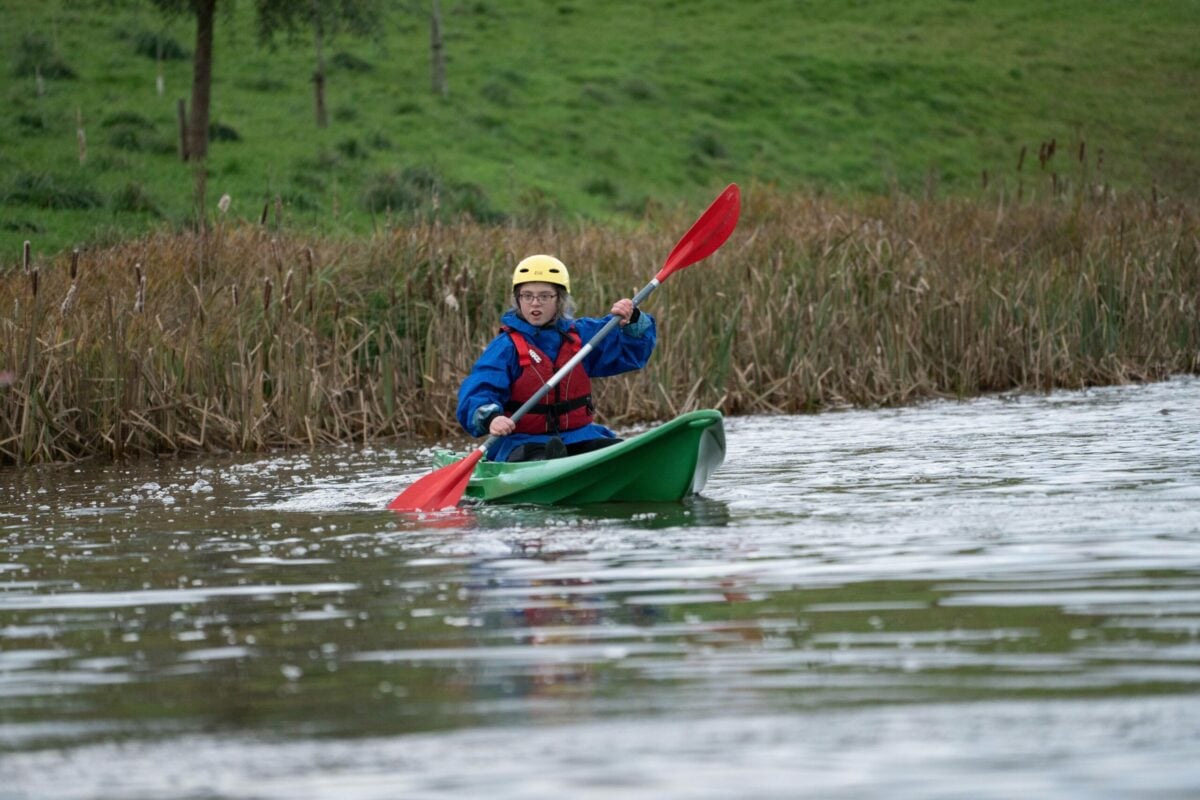 A young person kayaking, there is tall grass and a green hill behind them.
