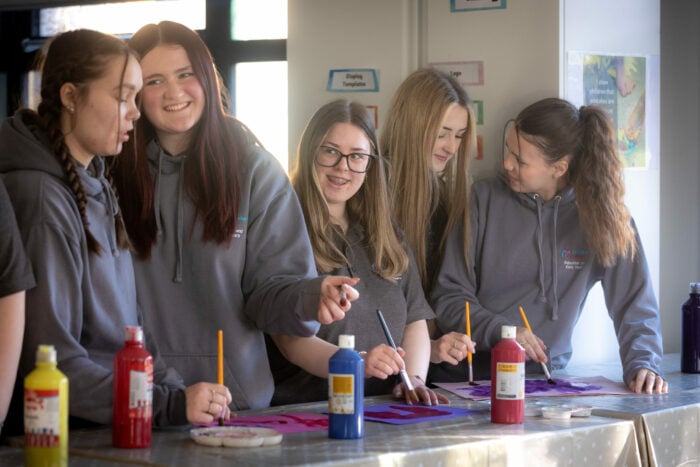 A group of five young women wearing gray hoodies are engaged in a painting activity. They are standing around a table covered with art supplies, including bottles of paint in red, yellow, and blue. The women are smiling and interacting with each other as they work on their projects, which appear to involve colorful designs. The background features a bright, well-lit room with art-related materials displayed on the walls.