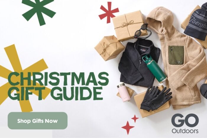 Flat lay of winter outdoor clothing and accessories arranged with wrapped gift boxes. Items include a beige fleece hoodie, black zip-up jacket, grey beanie hat with ‘Berghaus’ logo, black gloves, green insulated bottle, pink travel mug, and black slip-on shoes. Decorative green and red star shapes in the background. Text reads ‘Christmas Gift Guide’ with a green button labelled ‘Shop Gifts Now’ and the GO Outdoors logo.