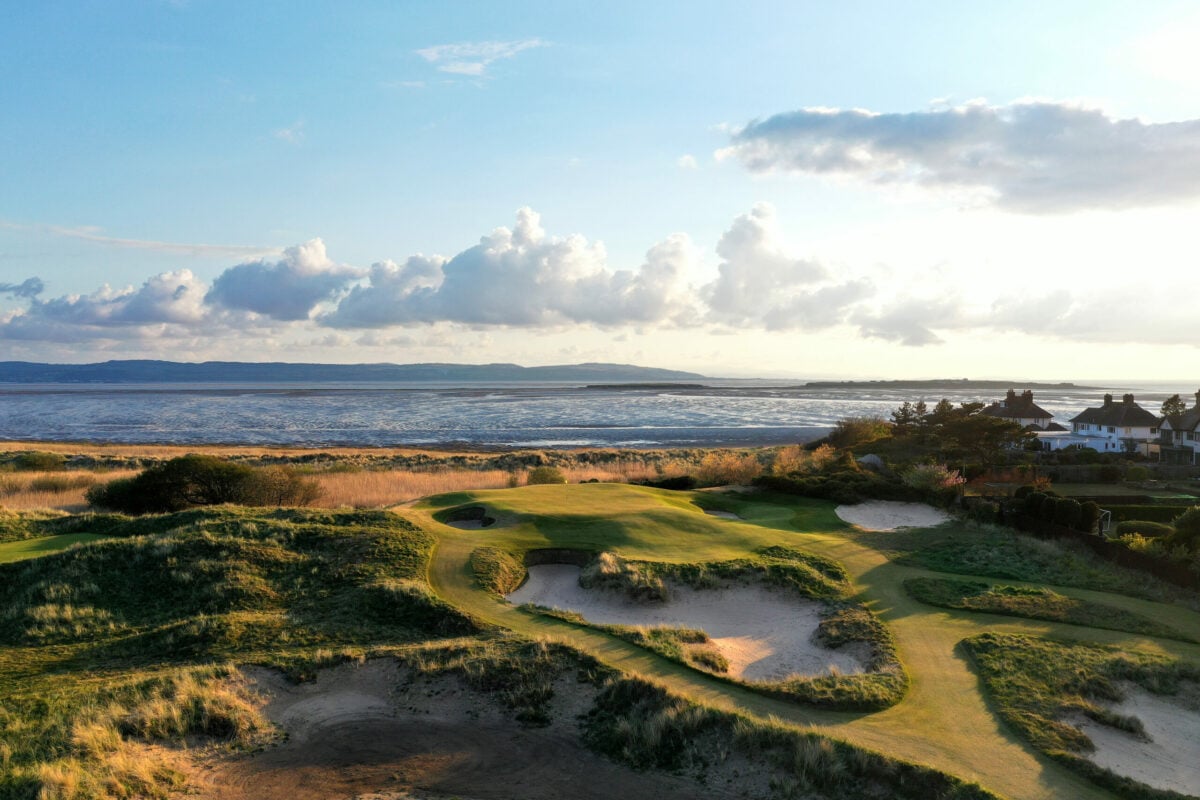 A scenic coastal golf course with rolling grassy dunes and sandy bunkers in the foreground, overlooking a calm sea under a partly cloudy sky. Houses are visible to the right, and distant hills stretch across the horizon.